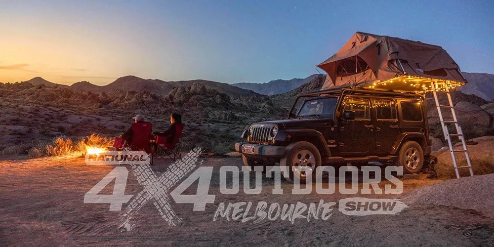 Promotional image for the National 4x4 Outdoors Show Melbourne featuring two people sitting by a campfire next to a Jeep with a rooftop tent and fairy lights, set in a rugged mountain landscape at sunset.