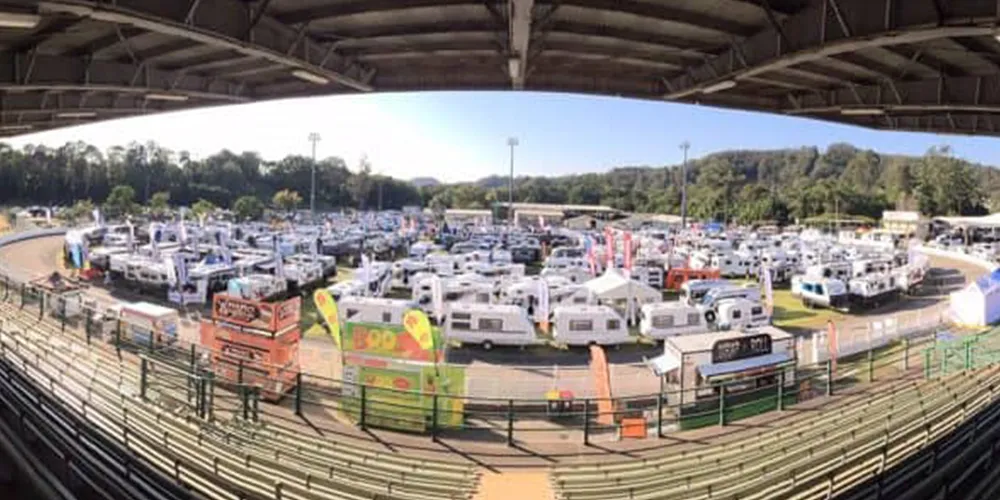 Wide-angle view of the Let’s Go Sunshine Coast Caravan & Outdoor Expo, showing numerous caravans, camper trailers, and exhibitor tents arranged around a showground track, viewed from under the grandstand roof.