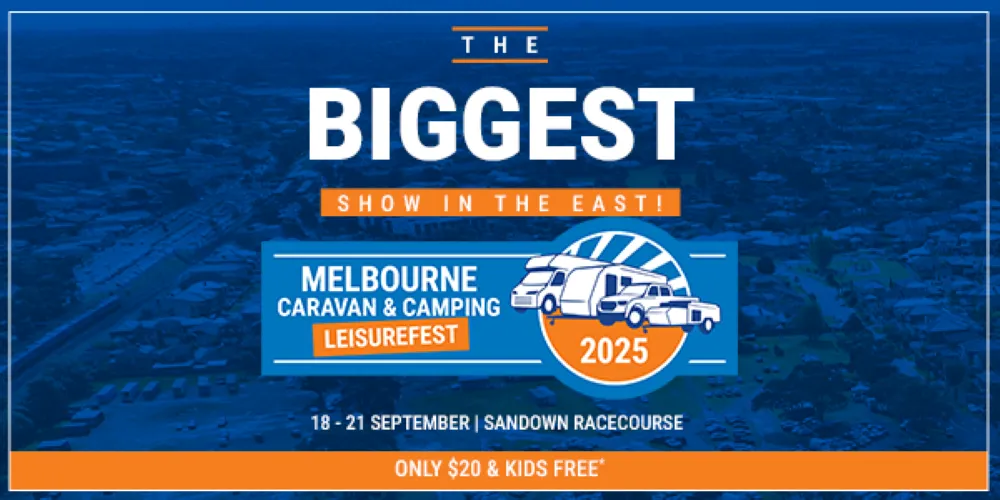 Crowds walk through rows of caravan displays at the Melbourne Caravan & Camping Leisurefest, with brand flags and touring vans lining both sides under a clear blue sky.