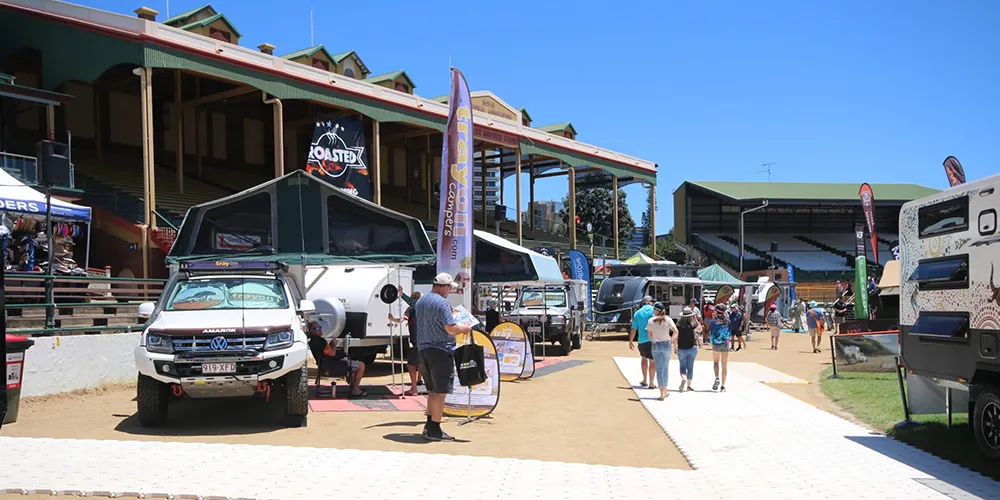 Visitors walk through outdoor displays of caravans and camper trailers at the Let’s Go Brisbane Caravan & Outdoor Sale, held at a showground with grandstands under clear blue skies.