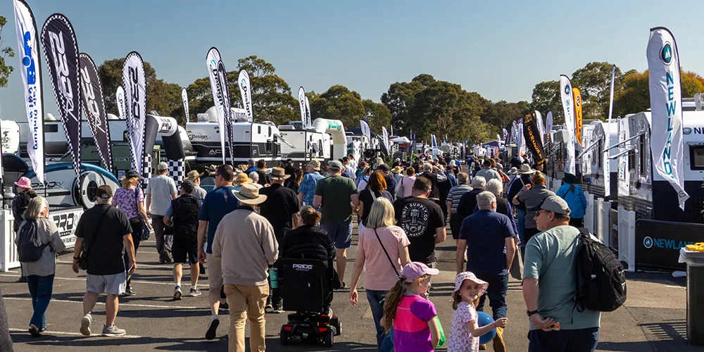 Crowds walk through rows of caravan displays at the Melbourne Caravan & Camping Leisurefest, with brand flags and touring vans lining both sides under a clear blue sky.