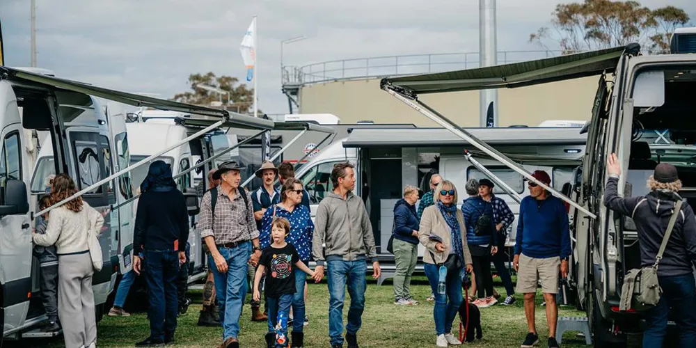 Aerial view of the The Busselton Caravan & Camping Show showing rows of caravans, camper trailers, and exhibitor tents set up on grassy grounds beside a racetrack, with trees and buildings in the background.